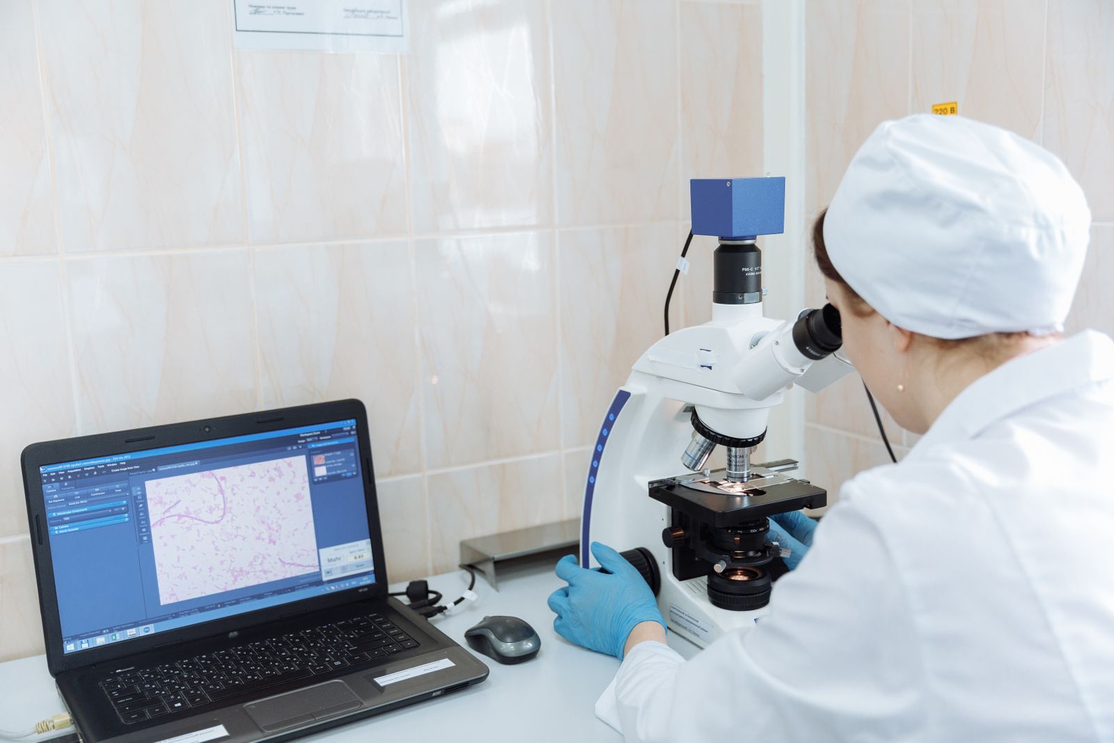 Scientist in a lab coat working at a microscope beside a laptop