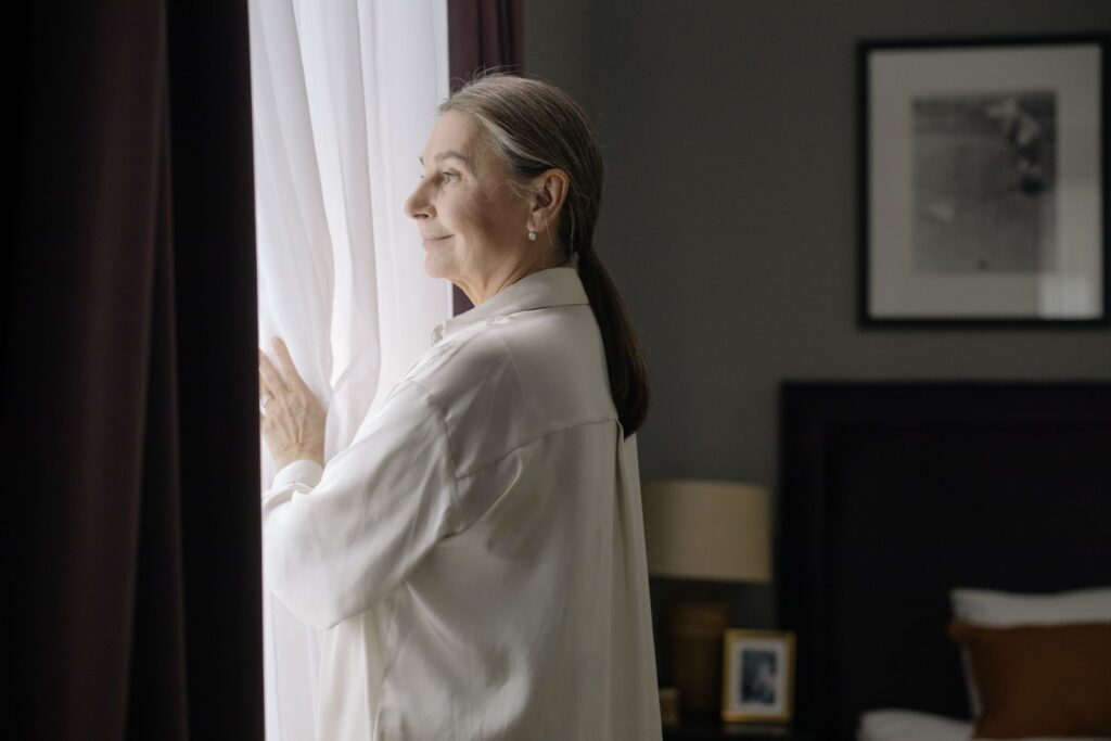Woman with grey hair standing thoughtfully at a window in soft daylight