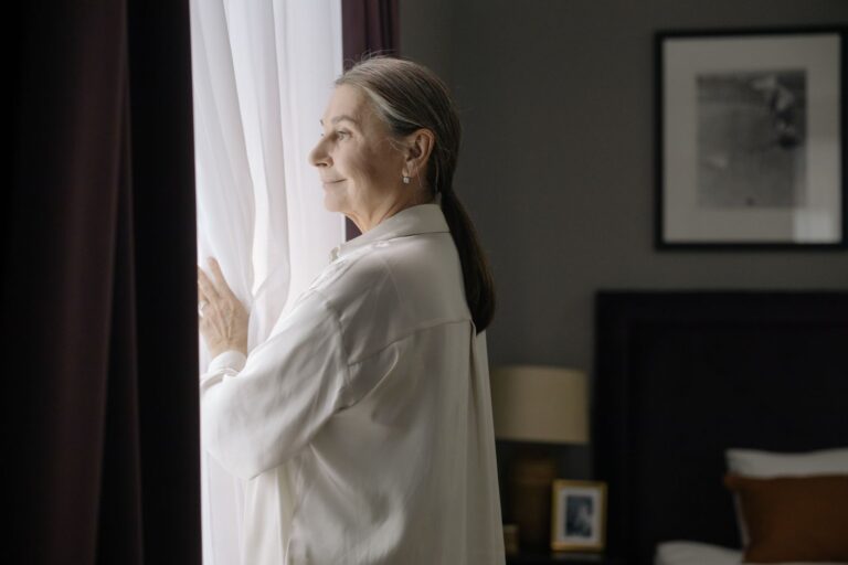 Woman with grey hair standing thoughtfully at a window in soft daylight