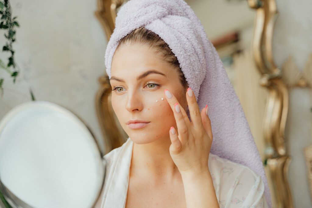 A woman with a hair towel applying moisturiser at a mirror